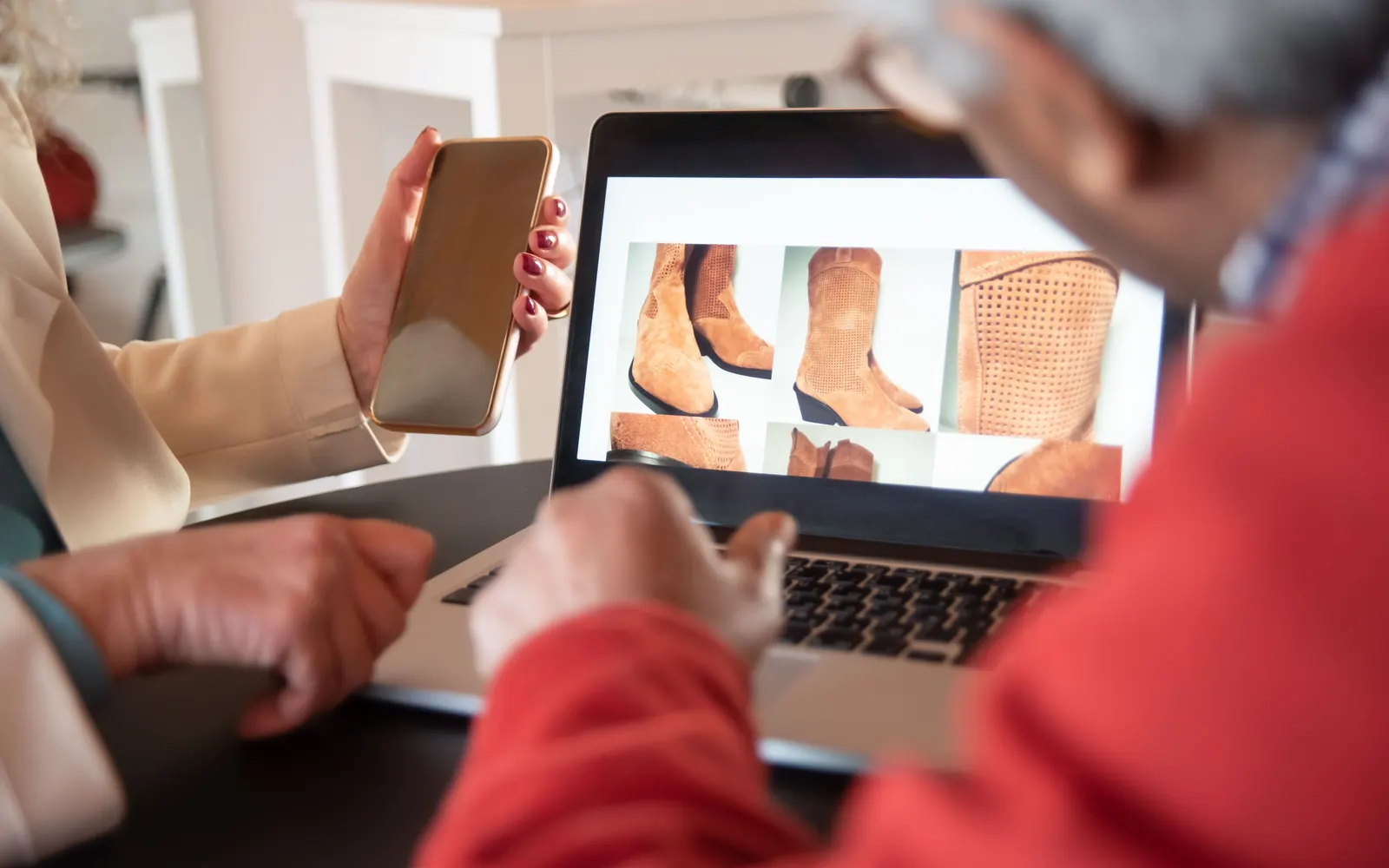 Clientes olhando fotos de botas na tela de um notebook enquanto um deles segura o celular